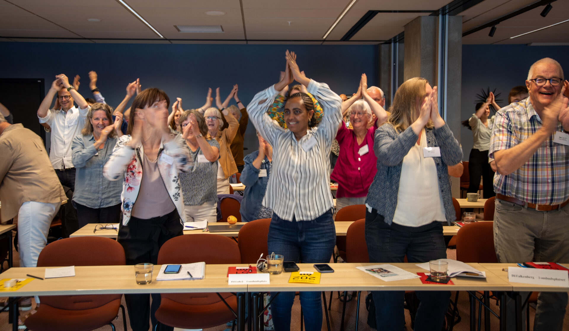 Ett stort antal människor i ett mötesrum har rest sig upp för att ta en danspaus framför stolarna.