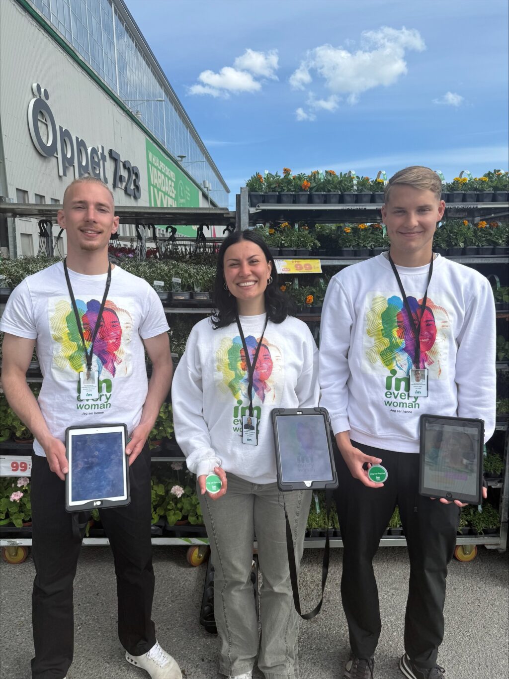 Två män och en kvinna står utanför en butik med skyltar och blomställningar, alla håller surfplattor och bär tröjor med texten "IM; every woman; Jag ser henne".